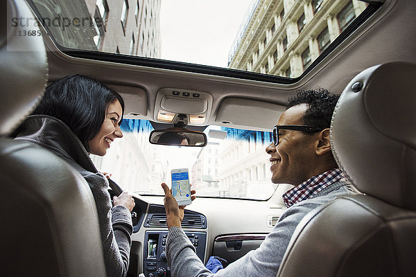Eine junge Frau und ein junger Mann in einem Auto  die vom Rücksitz aus eine Karte auf dem Display eines Mobiltelefons betrachten.