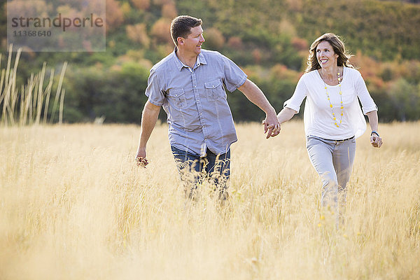 Lächelndes Paar  das sich die Hände hält  während es in einem Feld spazieren geht