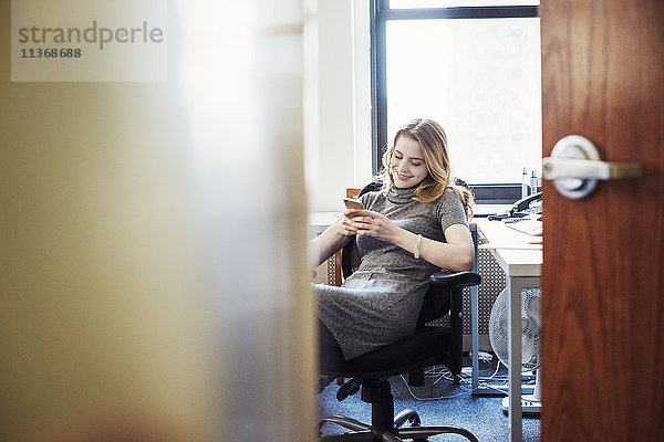 Eine Frau  die in einem Büro sitzt und ihr Telefon kontrolliert  durch eine offene Tür gesehen.