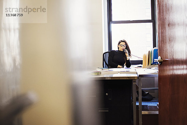 Eine junge Frau sitzt an einem Schreibtisch in einem Büro und hält sich ein Telefon an den Kopf.