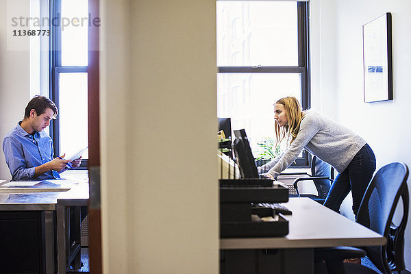 Die Aufnahme eines jungen Mannes  der in einem Büro sitzt und auf ein Mobiltelefon schaut  und einer jungen Frau  die in einem anderen Büro steht und an einem Computer arbeitet.
