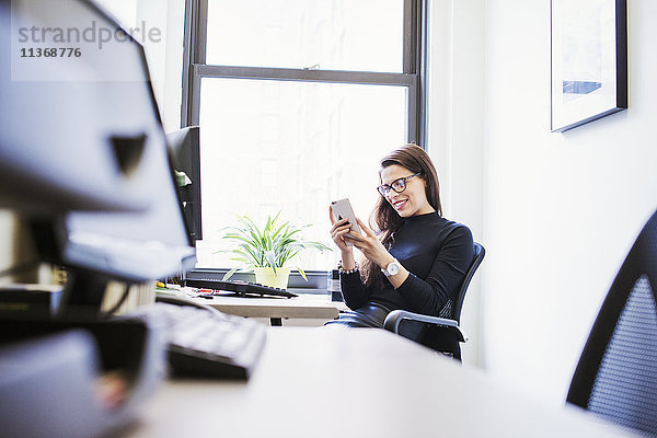 Eine junge Frau sitzt an einem Schreibtisch in einem Büro und schaut auf ein Handy.