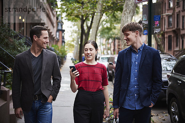 Zwei junge Männer und eine junge Frau gehen eine Straße in der Stadt entlang und schauen auf ein Handy.