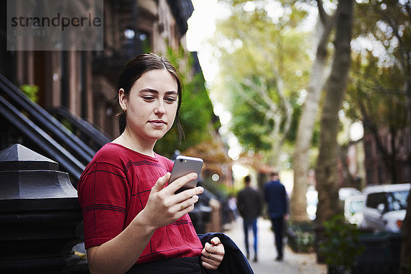 Eine junge Frau steht auf einer Straße in der Stadt und schaut auf ein Handy.