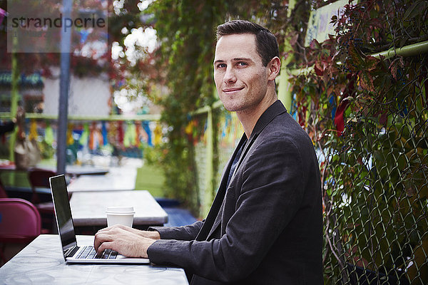 Ein junger Mann sitzt draußen an einem Tisch mit einem geöffneten Laptop und schaut ins Bild.