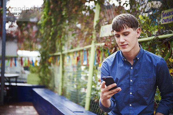 Ein junger Mann sitzt im Freien und schaut auf ein Handy.