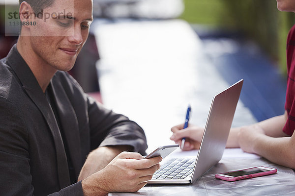 Ein junger Mann  der mit einem offenen Laptop an einem Tisch im Freien sitzt und auf ein Handy schaut.