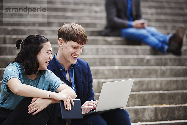 Eine junge Frau und ein junger Mann sitzen auf einer Freitreppe im Freien und schauen gemeinsam auf einen Laptop.