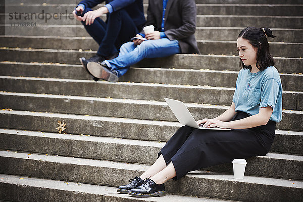 Eine junge Frau sitzt auf einer Freitreppe im Freien und arbeitet an einem Laptop  mit zwei Männern im Hintergrund.