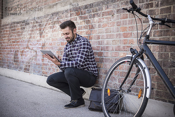 Junger Mann  der ein digitales Tablet benutzt und an einer Mauer sitzt