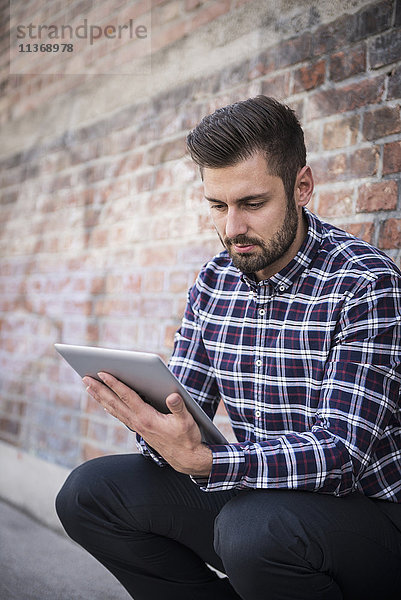 Junger Mann  der ein digitales Tablet benutzt und an einer Mauer sitzt
