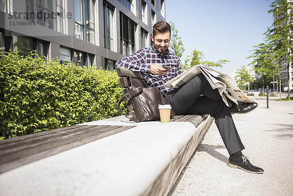 Junger Mann sitzt auf einer Bank und schreibt eine SMS auf seinem Smartphone