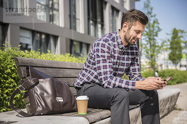Junger Mann sitzt auf einer Bank und schreibt eine SMS auf seinem Smartphone