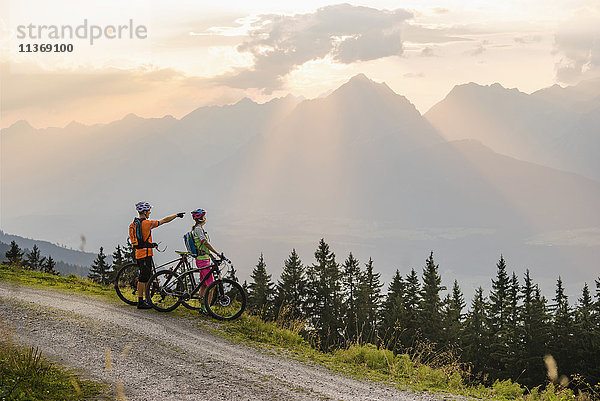 Junges Mountainbike-Paar steht auf einer unbefestigten Straße und betrachtet die Aussicht bei Sonnenuntergang  Zillertal  Tirol  Österreich