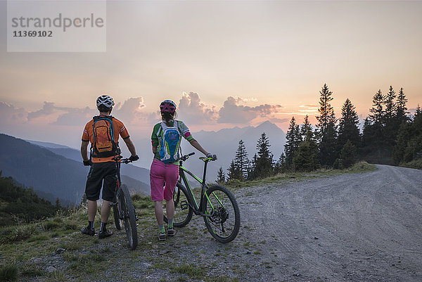 Rückansicht eines jungen Mountainbike-Paares  das in der alpinen Landschaft steht und die Aussicht bei Sonnenuntergang betrachtet  Zillertal  Tirol  Österreich