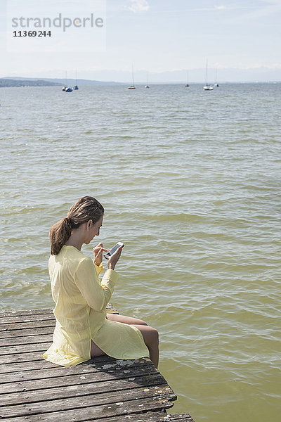 Frau benutzt Smartphone und sitzt auf Steg am See  Ammersee  Oberbayern  Deutschland