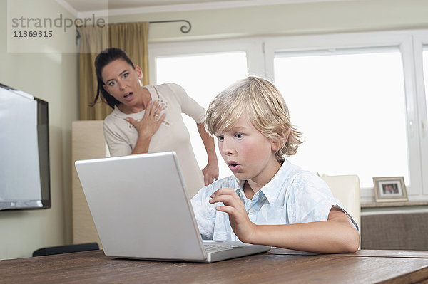 Schockierte Frau beobachtet ihren Sohn bei der Benutzung eines Laptops im Wohnzimmer