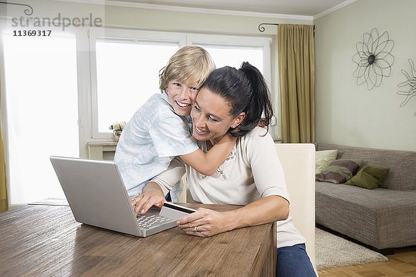 Junge  der seine Mutter umarmt  während sie auf dem Laptop online einkauft