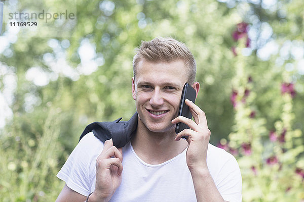 Porträt eines jungen Mannes  der in einem städtischen Garten mit einem Mobiltelefon spricht