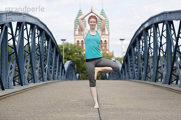 Junge Frau  die auf einer Brücke in einer Großstadt Yoga in Baumstellung macht