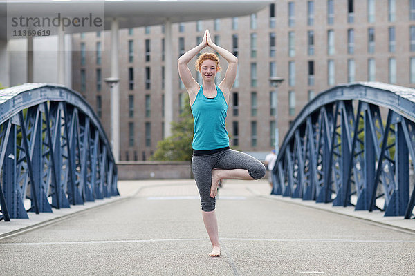 Junge Frau  die auf einer Brücke in einer Großstadt Yoga in Baumstellung macht