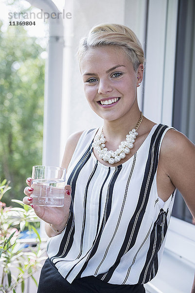 Porträt einer jungen Frau  die auf dem Balkon Wasser trinkt und lächelt