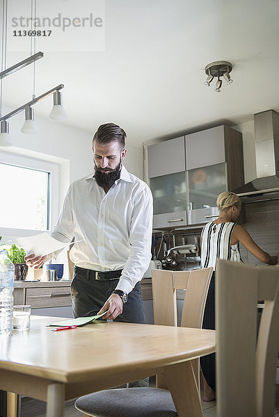 Junger Mann mit Dokumenten am Küchentisch und Frau bei der Arbeit im Hintergrund