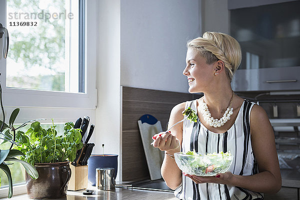 Junge Frau isst Salat in der Küche und lächelt