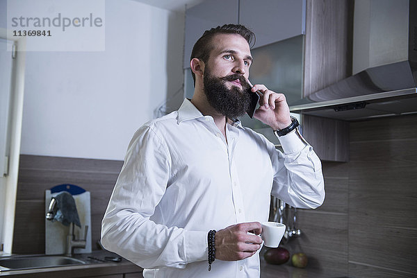 Junger Mann spricht mit seinem Smartphone und trinkt Kaffee in der Küche