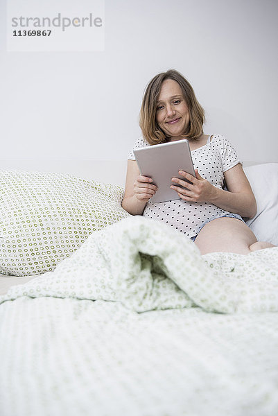 Glückliche schwangere Frau mit digitalem Tablet im Bett