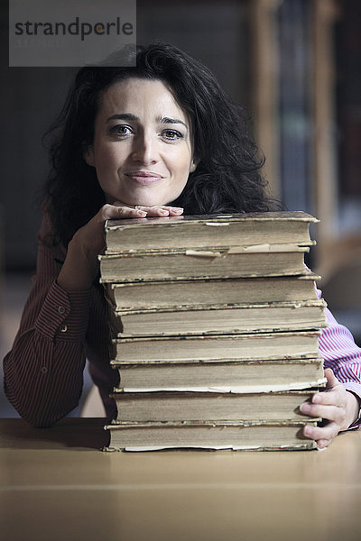 Braunhaarige junge Frau in einer Bibliothek  deren Kopf und eine Hand auf einem Stapel alter Bücher ruhen.