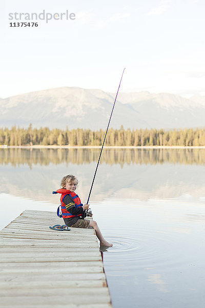 Kaukasischer Junge sitzt auf einem Steg am See und hält eine Angelrute