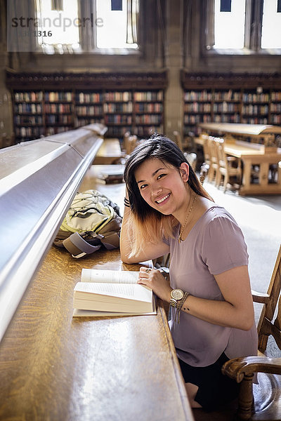 Lächelnde chinesische Frau sitzt in der Bibliothek und liest ein Buch