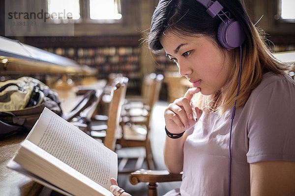 Chinesische Frau in der Bibliothek liest ein Buch und hört Kopfhörer