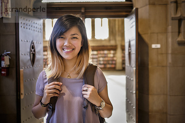 Chinesische Frau steht in der Bibliothek und trägt einen Rucksack