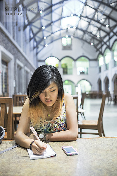 Chinesische Frau sitzt in der Bibliothek und schreibt in ein Notizbuch
