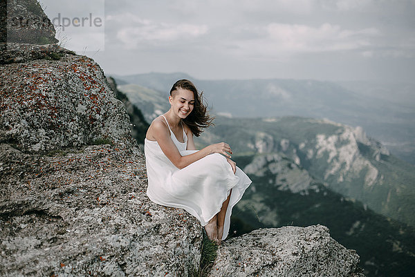 Lächelnde kaukasische Frau sitzt auf einem Felsen mit Blick auf die Landschaft