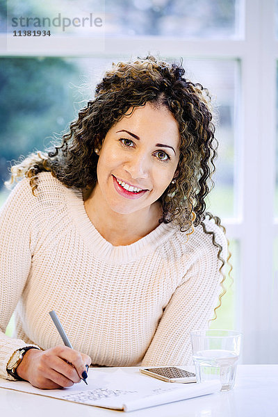 Lächelnde Frau sitzt am Tisch und schreibt mit einem Stift auf einen Notizblock