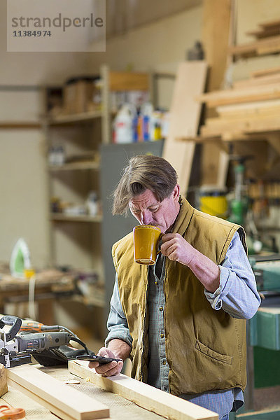 Kaukasischer Tischler trinkt Kaffee und schreibt eine SMS auf seinem Handy in der Werkstatt