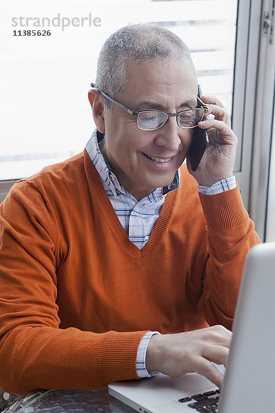 Lächelnder hispanischer Mann beim Multitasking mit Handy und Laptop