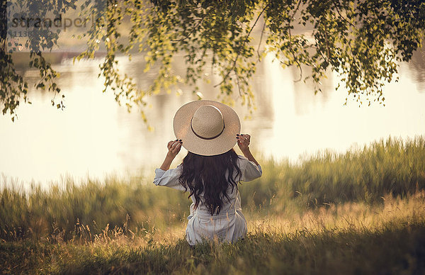 Kaukasische Frau sitzt im Gras in der Nähe des Flusses und hält einen Hut