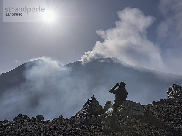 Bergwanderer schaut auf zwei rauchende Ostkrater im Gegenlicht  Blick vom Aussichtspunkt Rocca della Valle  Ätna  Vulkan  Nordostflanke  Sizilien  Italien  Europa