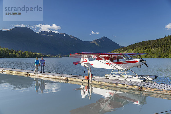 Eine Frau und ein Mann stehen auf einem Dock  an dem ein Wasserflugzeug angedockt ist  Trail Lake Float Plane Base  Moose Pass  Kenai Peninsula  Southcentral Alaska  USA