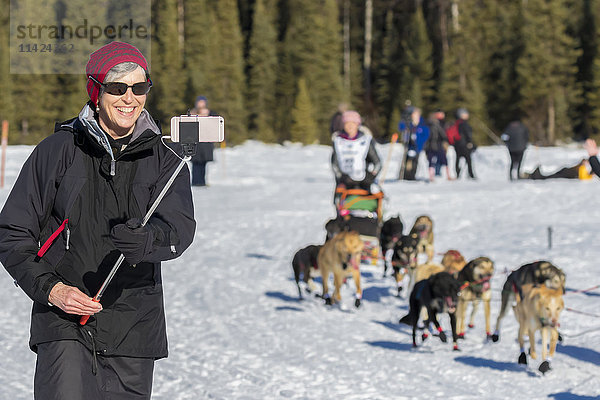 Eine Touristin macht ein Smartphone-Foto von einem Musher beim Start des Iditarod-Rennens am Willow Lake  Alaska  USA  Winter.
