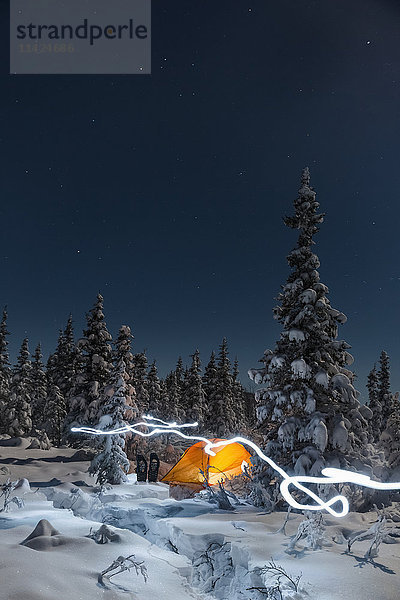 Eine Lichtspur führt an einem orangefarbenen Zelt inmitten eines verschneiten  immergrünen Waldes vorbei  Schneeschuhe vor dem Zelt  Mondlicht wirft Schatten in einer klaren Winternacht  Interior Alaska; Gakona  Alaska  Vereinigte Staaten von Amerika'.