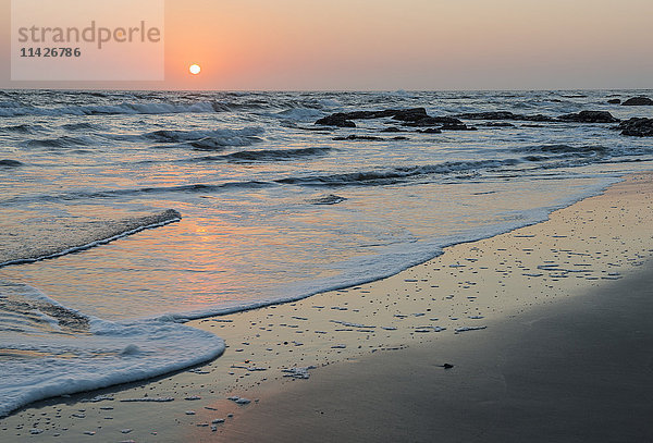 Friedlicher und ruhiger Sonnenuntergang über dem Atlantischen Ozean mit orange- und goldfarbener Sonne am Himmel  sandiger Küste und schäumender Brandung; Namibia'.