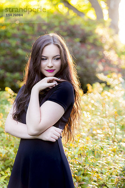 Ein junges Teenager-Mädchen in einem modischen schwarzen Kleid steht in einer nachdenklichen Pose in einem sonnigen Park; Vancouver  British Columbia  Kanada'.