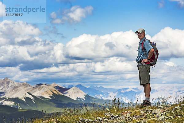 Männlicher Wanderer steht auf einem felsigen Hügel und überblickt Bergkette und Tal mit blauem Himmel und Wolken  westlich von Bragg Creek; Alberta  Kanada '