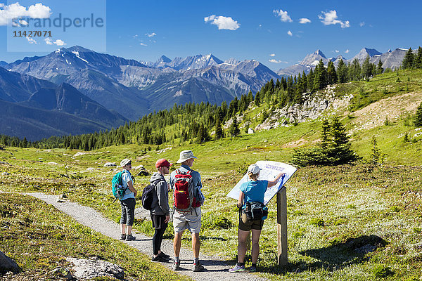 Eine Gruppe von Wanderern betrachtet ein Hinweisschild auf einem Wiesenweg mit einer Bergkette in der Ferne und blauem Himmel und Wolken; Banff  Alberta  Kanada'.