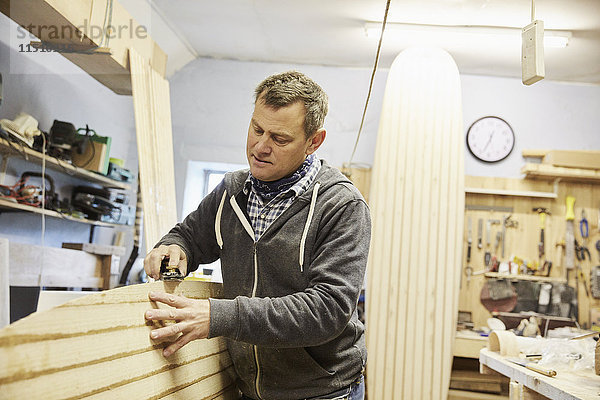 Mann  der in einer Werkstatt steht und die Oberfläche eines hölzernen Surfbretts mit einem Handwerkzeug glättet und formt.